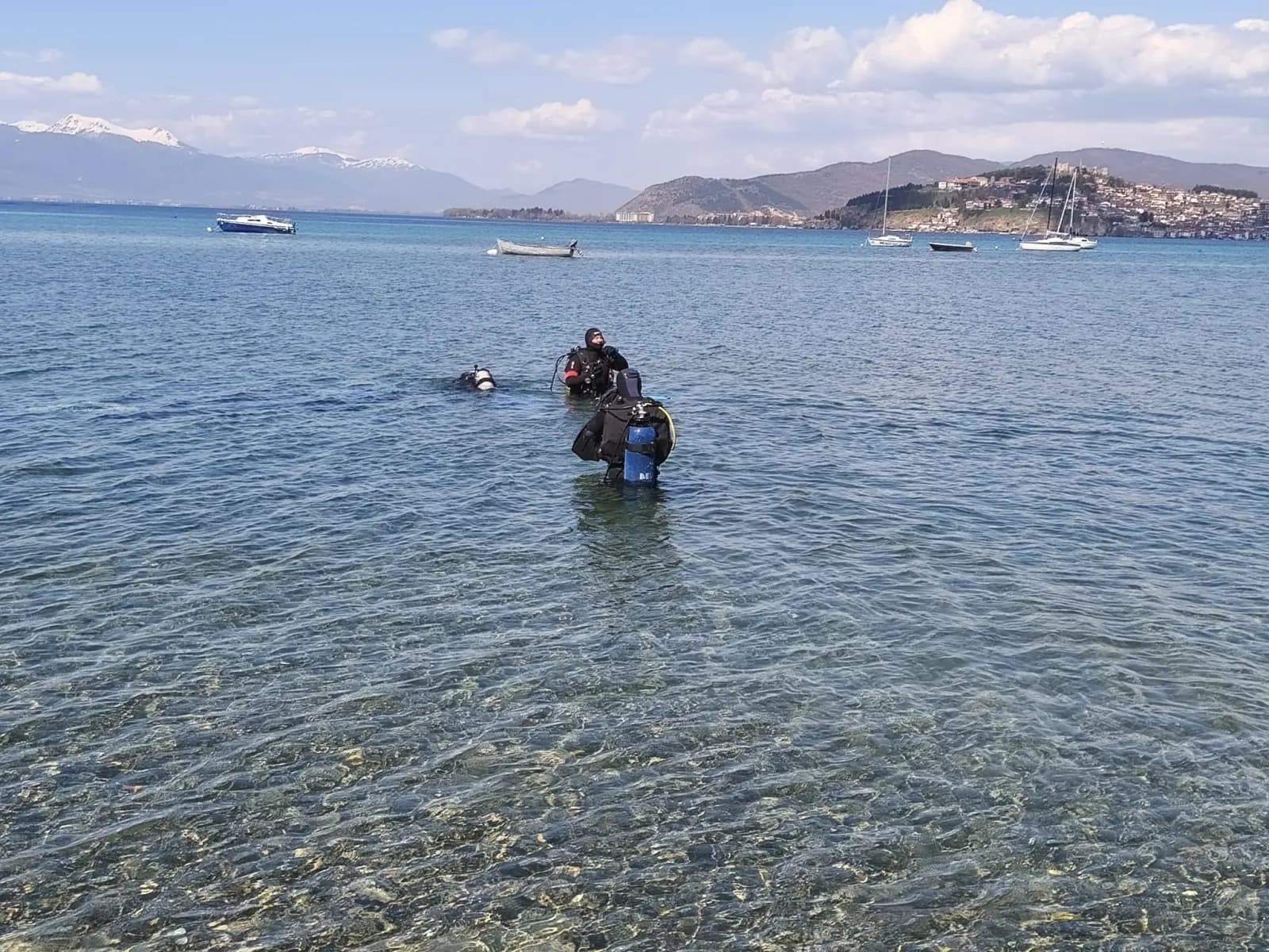 Two scuba divers in shallow water at Kaj Divono beach with boats and the city of Ohrid in the background
