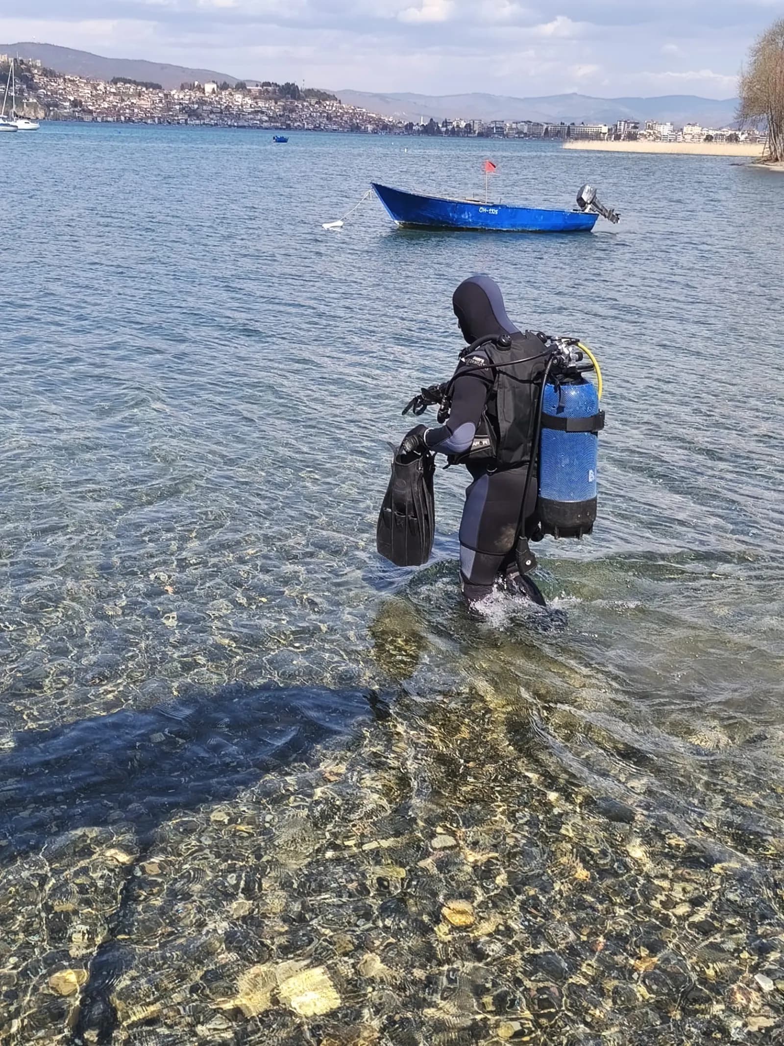A scuba diver walking out of Lake Ohrid carrying full diving gear, with the Old Town of Ohrid in view