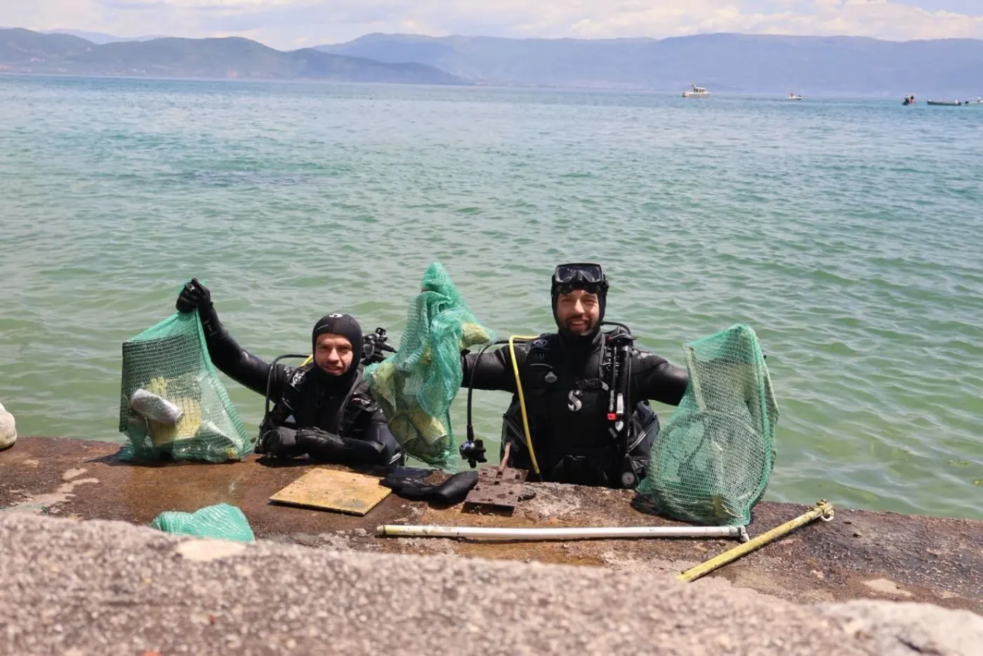 Two scuba divers on the shore holding mesh bags full of glass bottles and metal debris collected from the lakebed